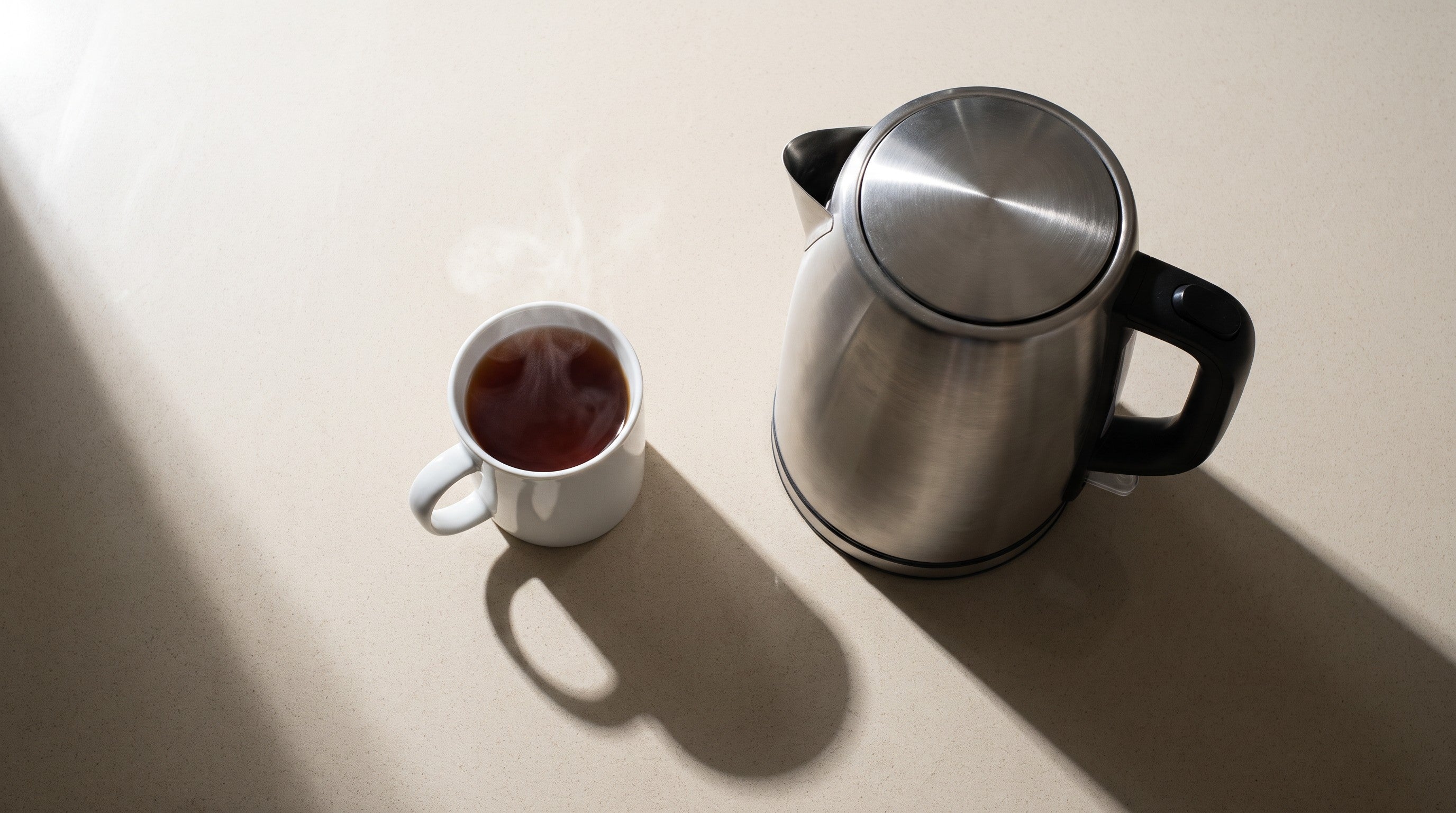 Image of Overhead view of a single small mug of tea beside a full-size stainless steel kettle, showing the difference in size between one cup and a full kettle