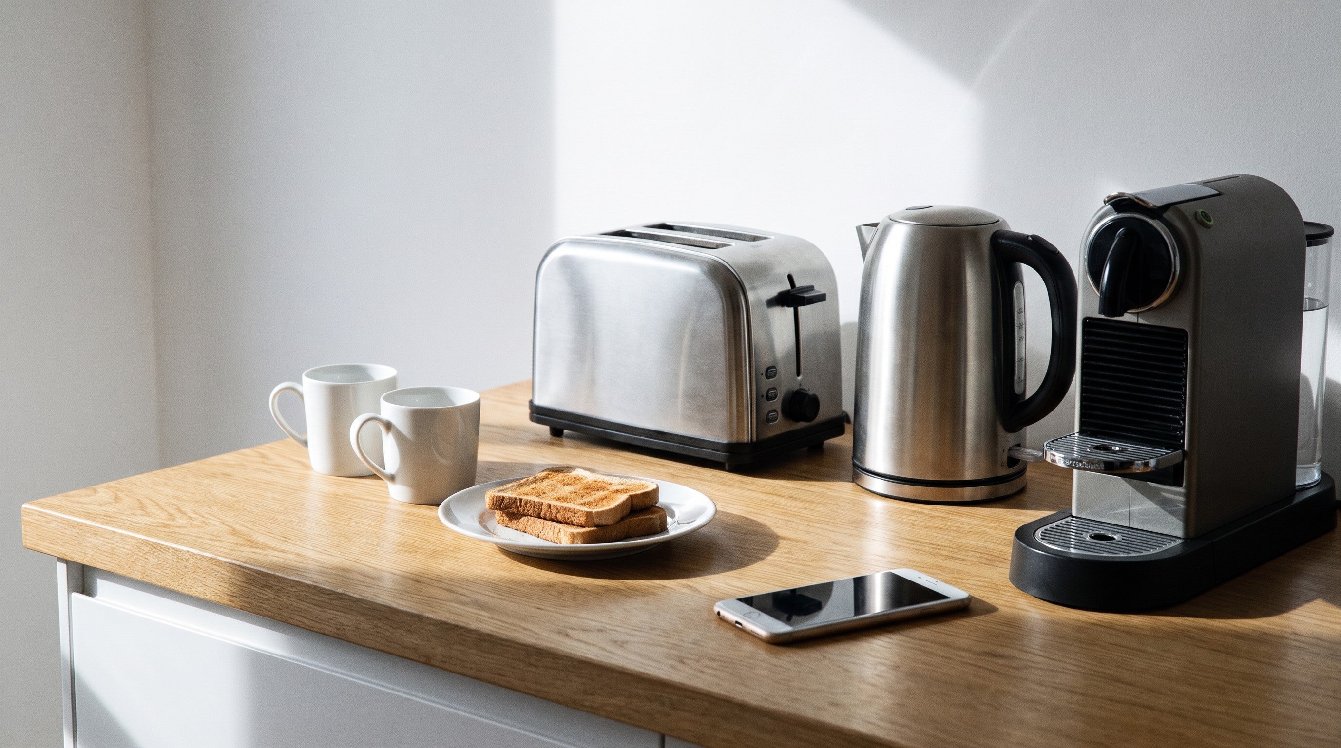 Image of A toaster, electric kettle and coffee machine lined up on a wooden worktop at breakfast, with two mugs, a plate of toast and a phone lying face up