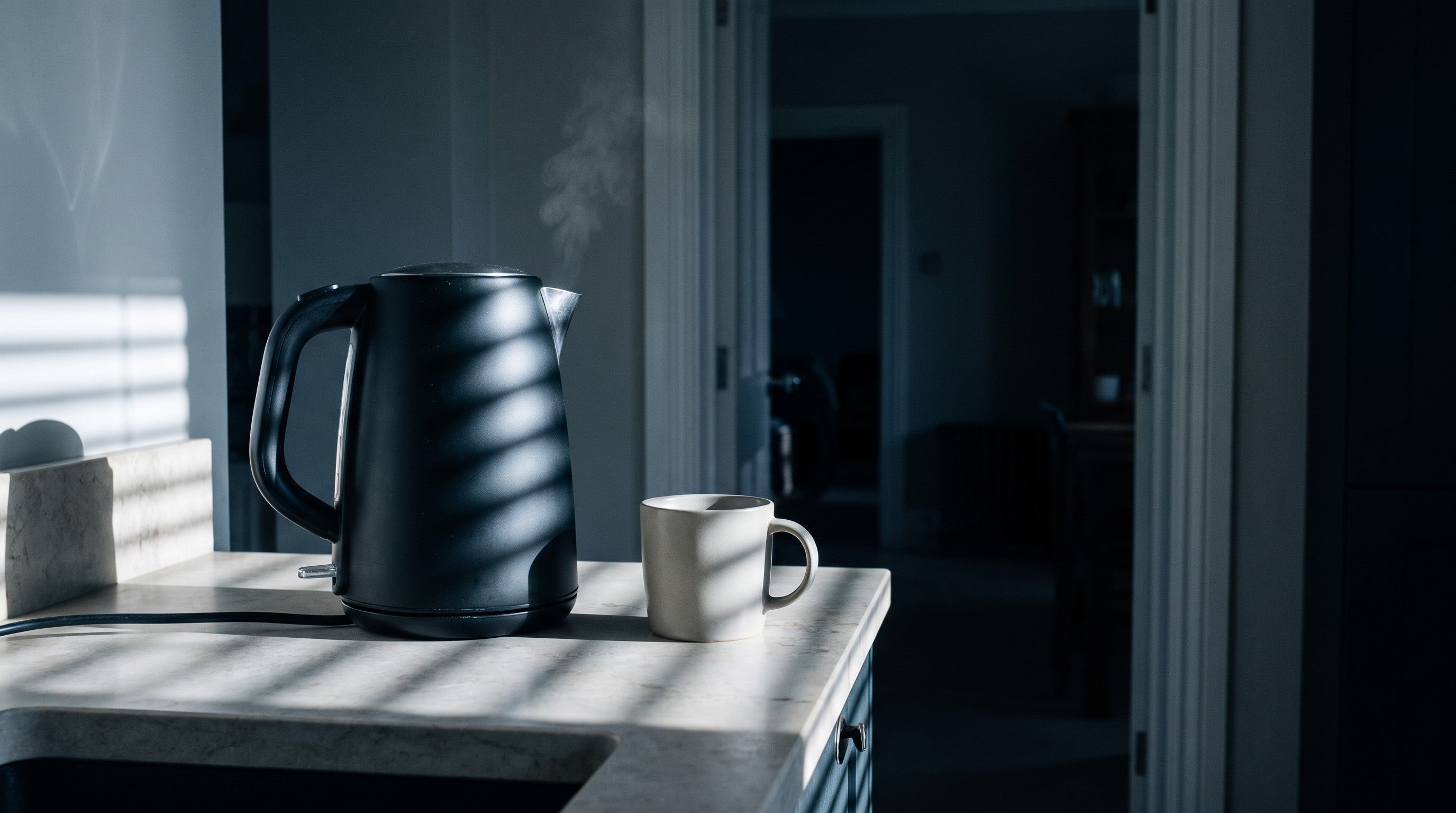 Image of A matt black electric kettle on a worktop in dim early morning light with a single mug beside it and the rest of the house dark and still behind