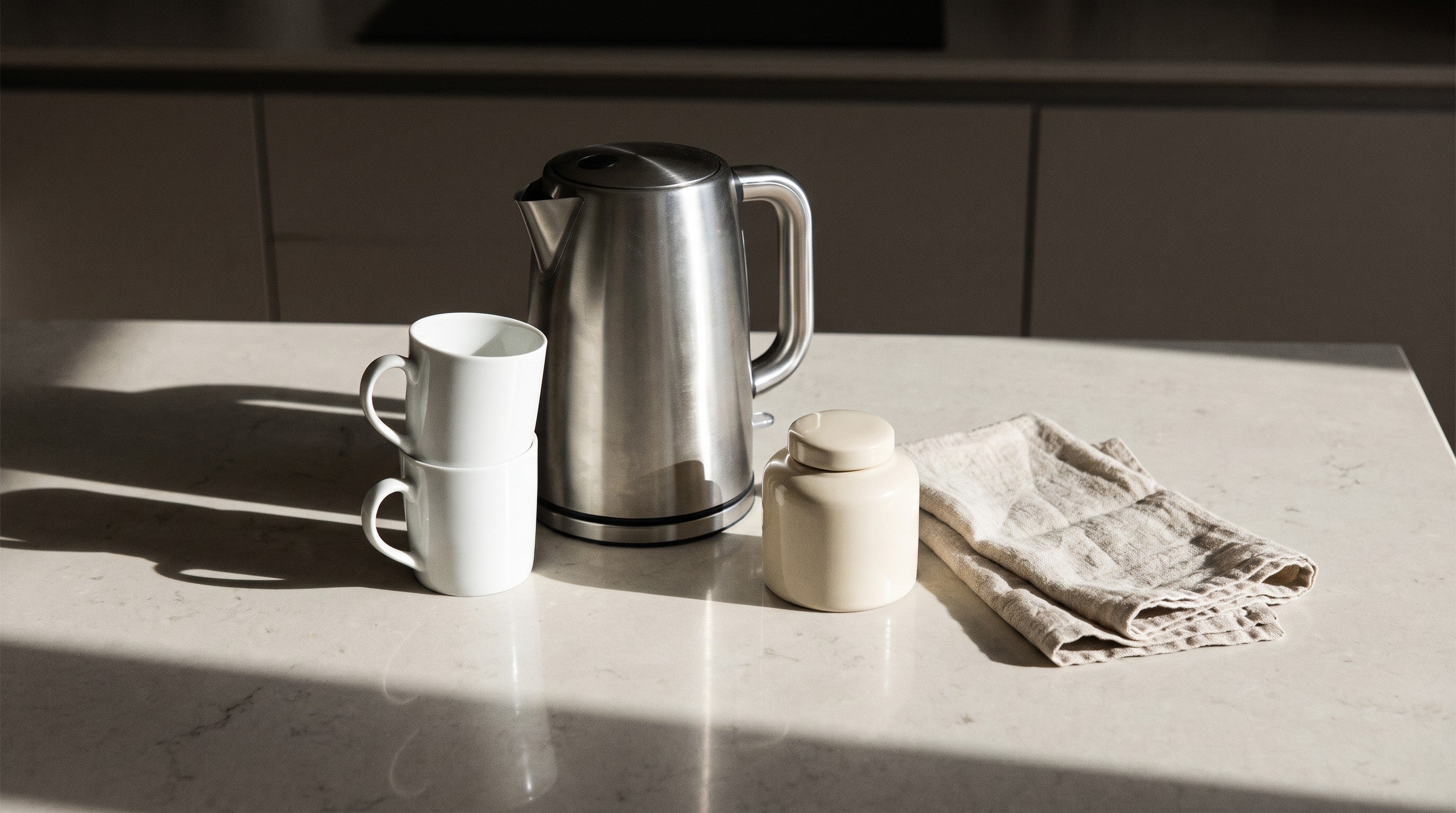 Image of A stainless steel kettle on a pale stone worktop in morning sunlight, beside two stacked white mugs, a cream tea caddy and a folded linen tea towel