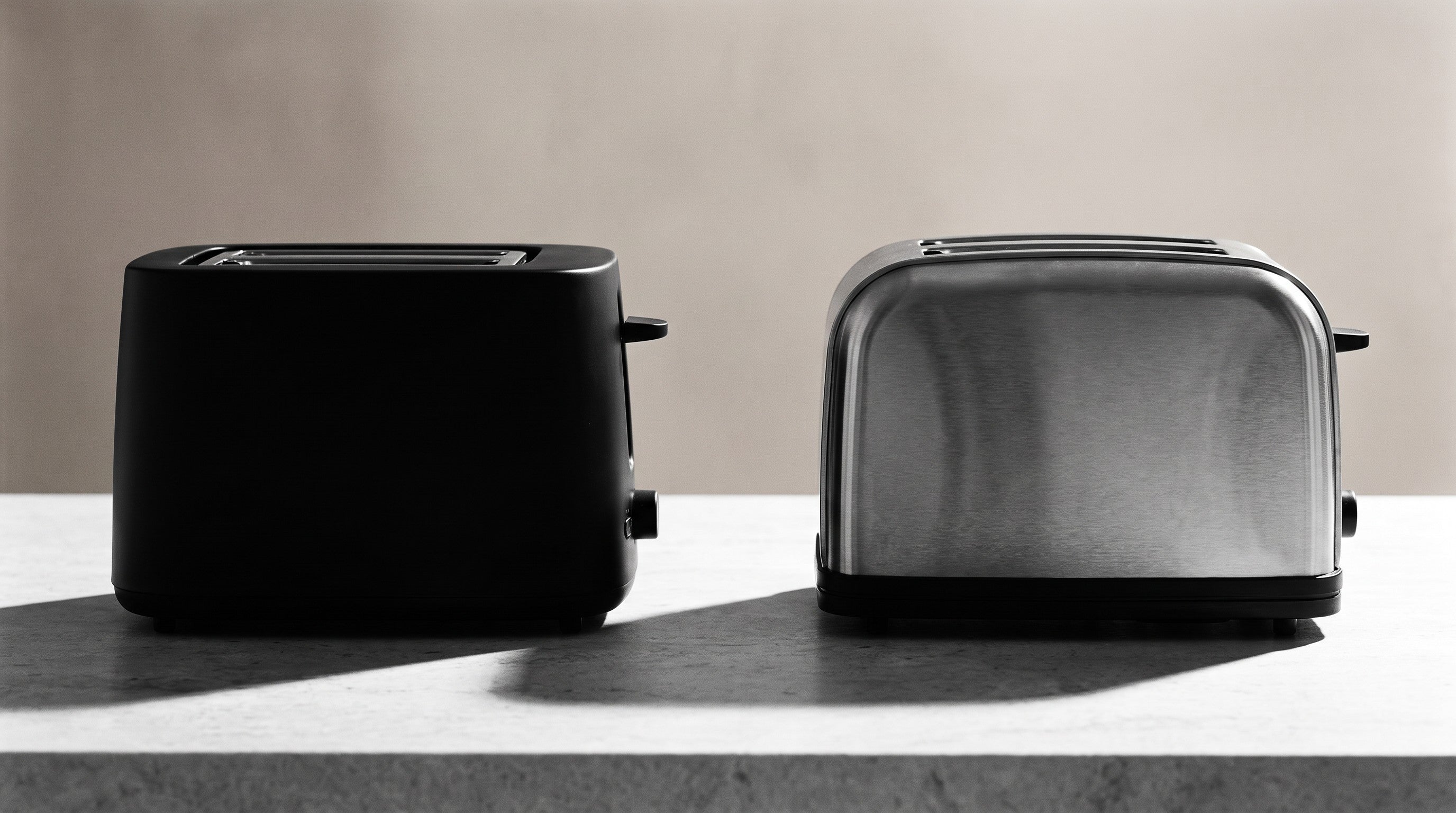 Image of A matt black toaster and a brushed stainless steel toaster side by side on a stone worktop, shown at the same scale for comparison