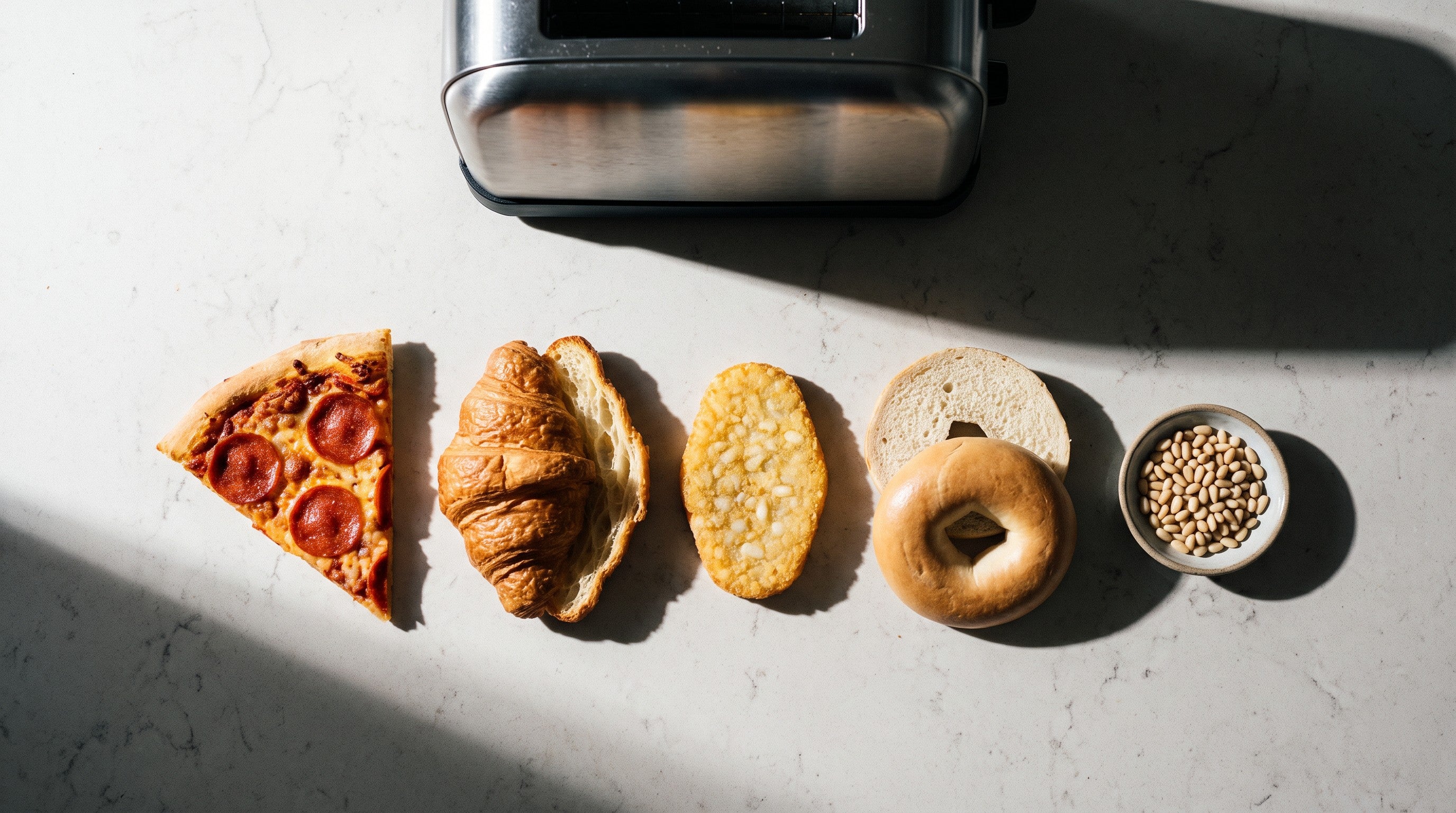 Image of A slice of pepperoni pizza, a halved croissant, a hash brown, a halved bagel and a small dish of pine nuts laid in a row on a worktop beside a toaster