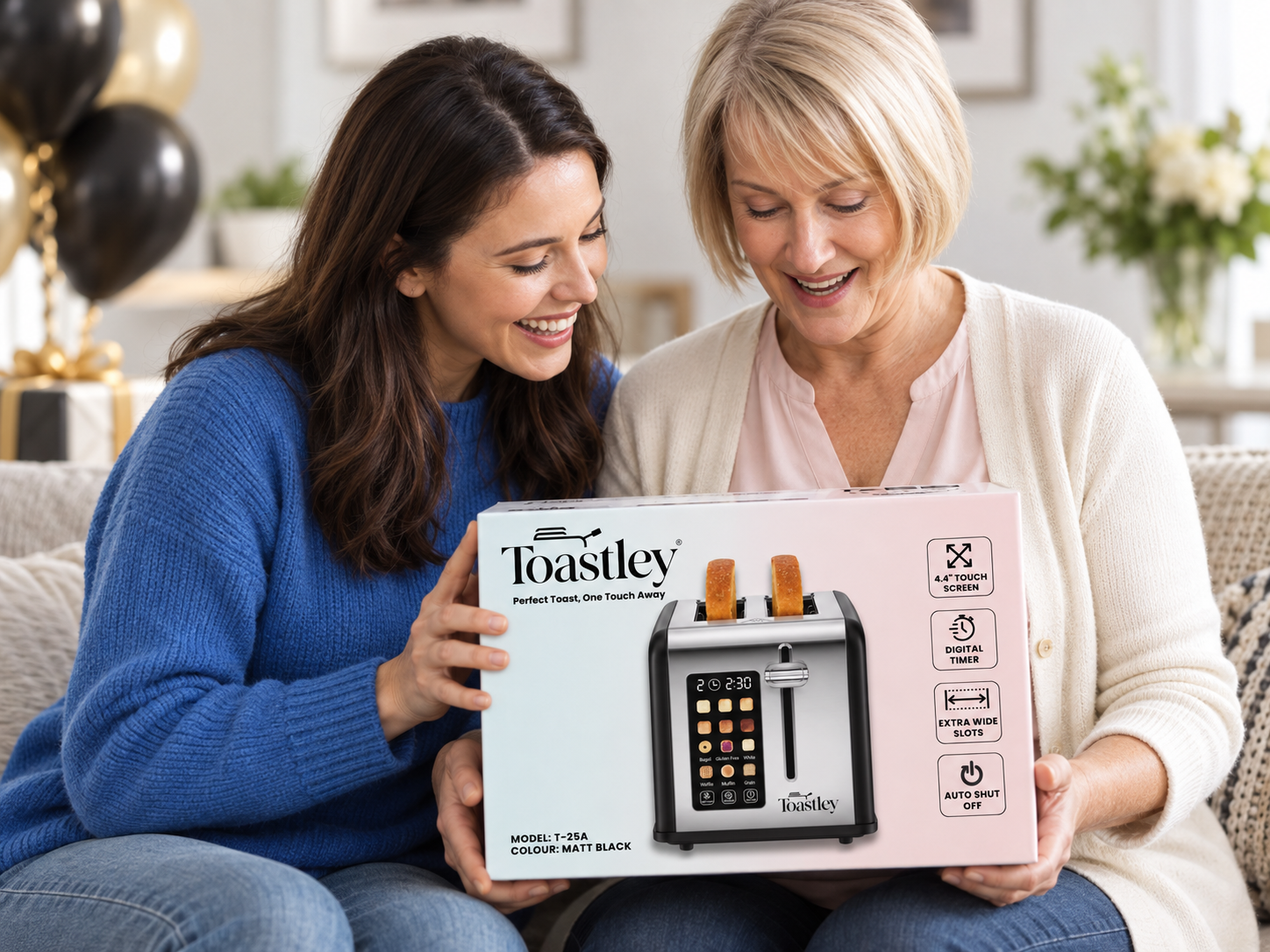 Image of Two women on a sofa with balloons behind them, smiling at a boxed Toastley 2 slice smart toaster in matt black being opened as a gift